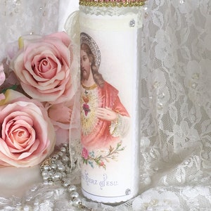 May include: A tall, white candle with an image of Jesus Christ, adorned with a gold and pink beaded rim and a lace ribbon. The candle features a decorative image and is embellished with rhinestones. A rosary and pink roses are in the background.