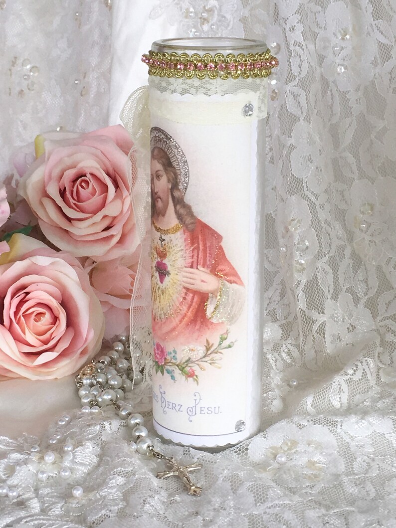 May include: A tall, white candle with an image of Jesus Christ, adorned with a gold and pink beaded rim and a lace ribbon. The candle features a decorative image and is embellished with rhinestones. A rosary and pink roses are in the background.