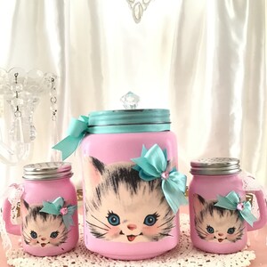 Puede incluir: Conjunto de tres tarros de vidrio rosa con un dise&ntilde;o de gatito vintage. Los tarros tienen tapas plateadas y est&aacute;n decorados con lazos azules y rosas. El tarro m&aacute;s grande tiene un pomo de cristal transparente en la tapa.