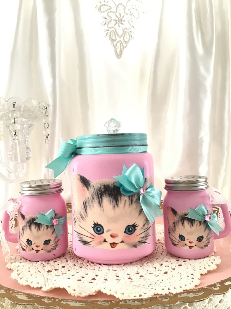 Puede incluir: Conjunto de tres tarros de vidrio rosa con un dise&ntilde;o de gatito vintage. Los tarros tienen tapas plateadas y est&aacute;n decorados con lazos azules y rosas. El tarro m&aacute;s grande tiene un pomo de cristal transparente en la tapa.