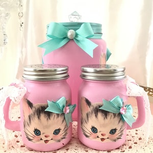 Puede incluir: Conjunto de tres tarros de vidrio rosa con caras de gatitos y lazos turquesa. Los tarros tienen tapas plateadas y est&aacute;n decorados con un borde de encaje blanco.