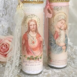 May include: Two decorative glass candle holders with religious imagery. One features Jesus, the other the Virgin Mary and baby Jesus. Both are adorned with lace, ribbons, and jeweled trim. The first candle holder has the text "Sacr&eacute; Coeur de J&eacute;sus, Ayez piti&eacute; de nous."