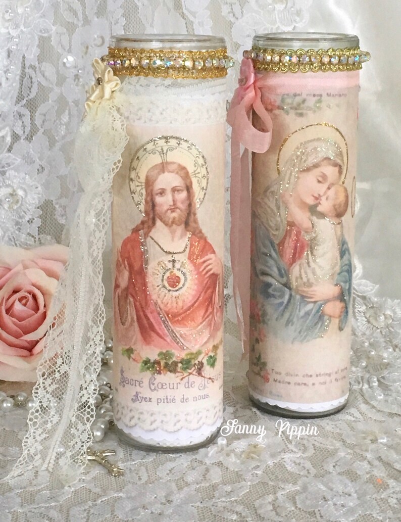 May include: Two decorative glass candle holders with religious imagery. One features Jesus, the other the Virgin Mary and baby Jesus. Both are adorned with lace, ribbons, and jeweled trim. The first candle holder has the text "Sacr&eacute; Coeur de J&eacute;sus, Ayez piti&eacute; de nous."