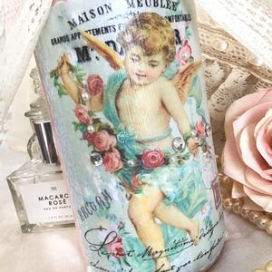 May include: A decorative bottle with a pink base and a vintage-style design. The bottle features an illustration of a cherub with roses, pearls, and the text "MAISON MEUBLEE". The bottle is embellished with rhinestones and floral accents.