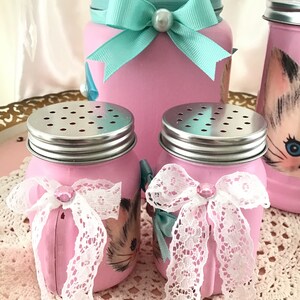 Puede incluir: Conjunto de tres tarros de vidrio rosa con lazos de encaje blanco y tapas plateadas. Los tarros est&aacute;n decorados con una ilustraci&oacute;n de gato vintage. El tarro m&aacute;s grande tiene una tapa turquesa con un pomo de cristal y un lazo turquesa.