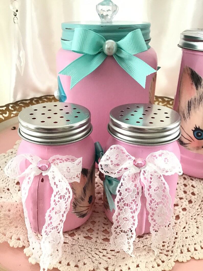 Puede incluir: Conjunto de tres tarros de vidrio rosa con lazos de encaje blanco y tapas plateadas. Los tarros est&aacute;n decorados con una ilustraci&oacute;n de gato vintage. El tarro m&aacute;s grande tiene una tapa turquesa con un pomo de cristal y un lazo turquesa.