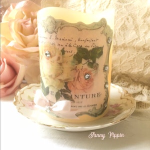 May include: A white candle with a vintage floral design and rhinestones. The candle is on a white saucer with a gold rim.