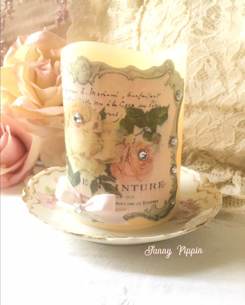 May include: A white candle with a vintage floral design and rhinestones. The candle is on a white saucer with a gold rim.