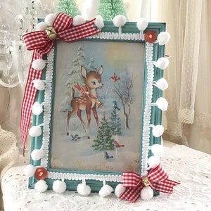 Puede incluir: Un marco de fotos turquesa con una escena invernal vintage con un ciervo, p&aacute;jaros y un &aacute;rbol de Navidad. El marco est&aacute; decorado con pompones blancos, lazos de cuadros rojos y blancos y peque&ntilde;as campanas doradas. El marco tiene las palabras "Christmas Cheer".
