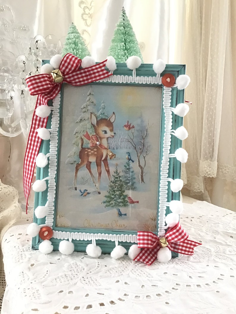 Puede incluir: Un marco de fotos turquesa con una escena invernal vintage con un ciervo, p&aacute;jaros y un &aacute;rbol de Navidad. El marco est&aacute; decorado con pompones blancos, lazos de cuadros rojos y blancos y peque&ntilde;as campanas doradas. El marco tiene las palabras "Christmas Cheer".