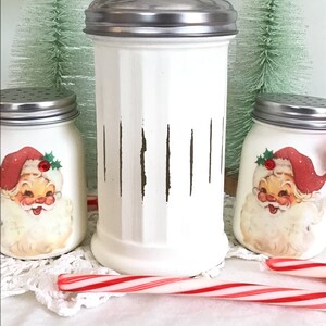 May include: Three white ceramic jars with vintage Santa Claus images and a silver lid. The jars are decorated with brown vertical stripes. A red and white candy cane is in the foreground.