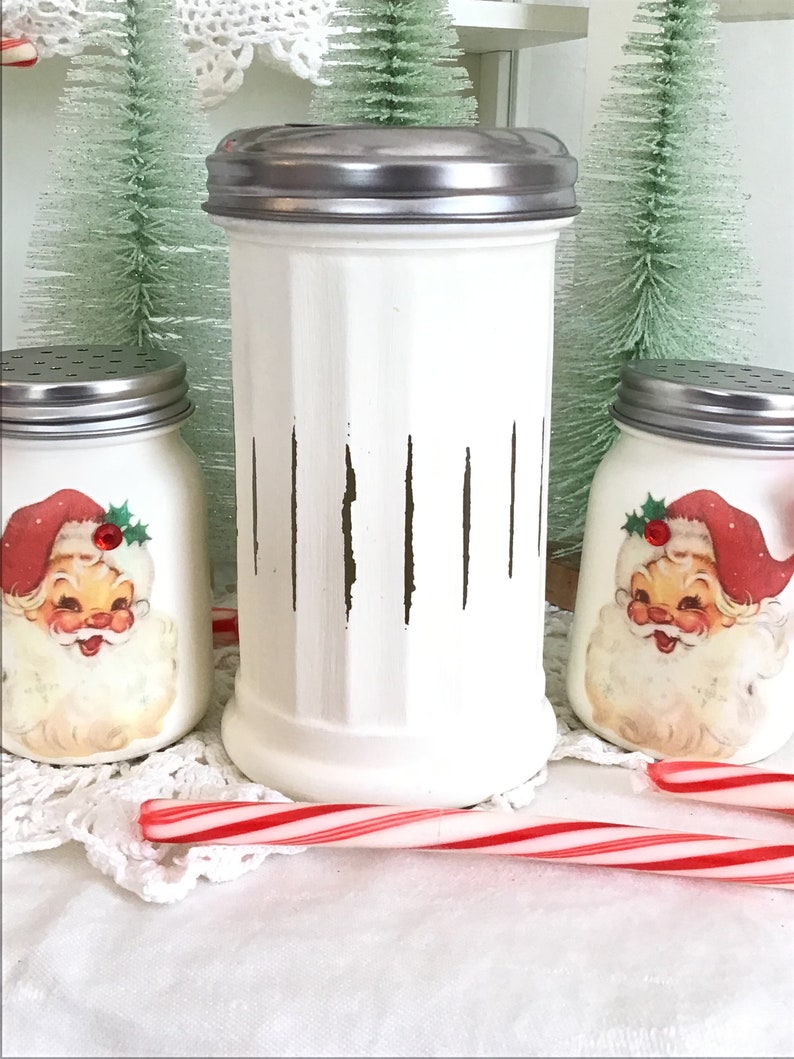May include: Three white ceramic jars with vintage Santa Claus images and a silver lid. The jars are decorated with brown vertical stripes. A red and white candy cane is in the foreground.