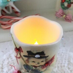 May include: A white ceramic candle holder with a snowman design. The candle is lit and glows yellow. The holder has a red and white ribbon and a bell.