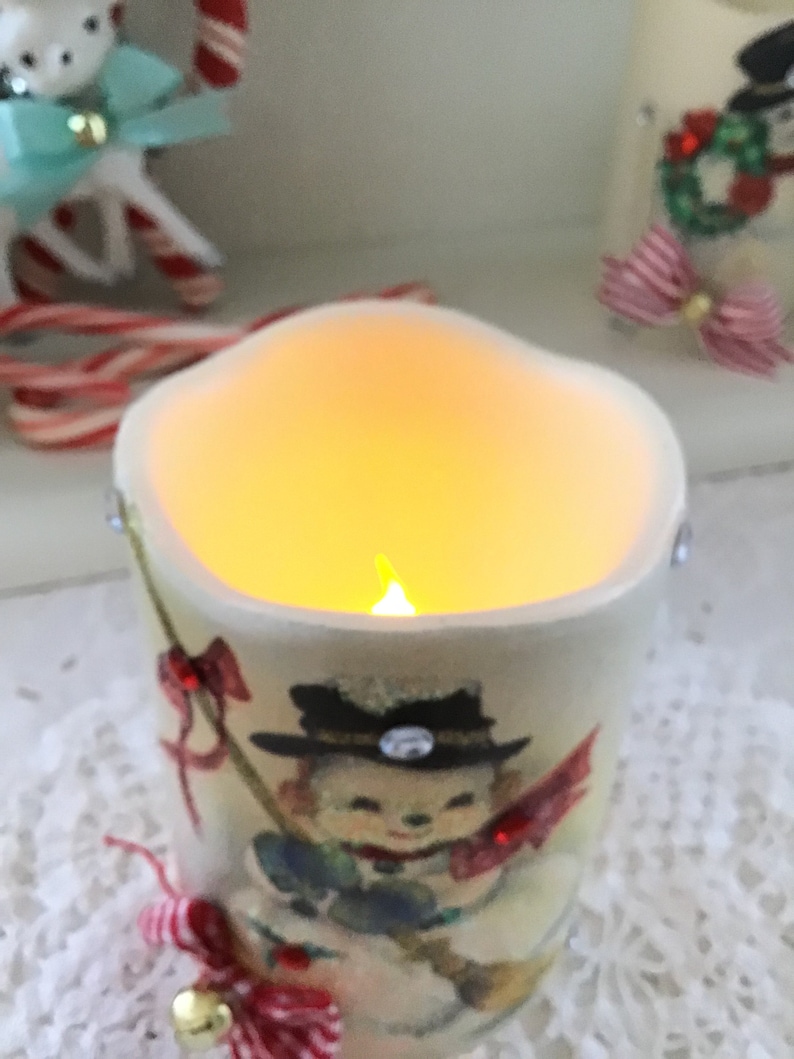 May include: A white ceramic candle holder with a snowman design. The candle is lit and glows yellow. The holder has a red and white ribbon and a bell.