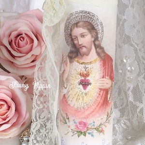 May include: A decorative glass candle holder featuring an image of Jesus with a halo and a heart. The candle holder is adorned with lace, pink roses, and a gold and pink trim. The text "Heiliges Herz Jesu" is visible.