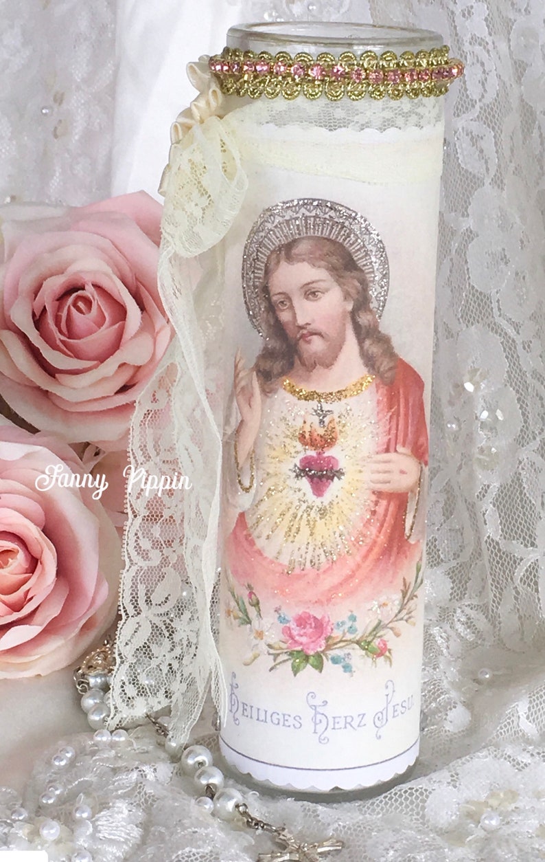 May include: A decorative glass candle holder featuring an image of Jesus with a halo and a heart. The candle holder is adorned with lace, pink roses, and a gold and pink trim. The text "Heiliges Herz Jesu" is visible.