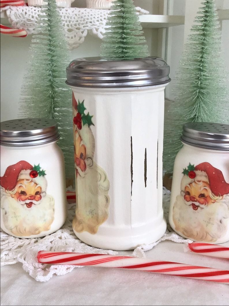 May include: Three white ceramic salt and pepper shakers with vintage Santa Claus illustrations and red holly accents. The shakers have silver lids and are sitting on a white doily with candy canes.