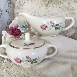 May include: A white creamer and sugar set with pink rose floral designs. The creamer has a curved spout and a handle. The sugar bowl has a lid with a gold trim.