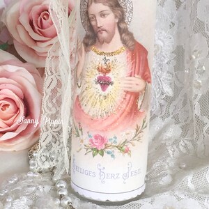 May include: A white candle featuring an image of Jesus with a heart and halo. The candle has the words "Heiliges Herz Jesu" and is decorated with pink roses and lace.