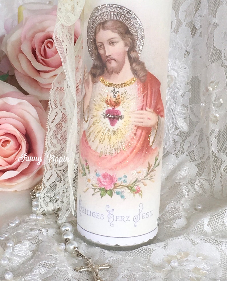 May include: A white candle featuring an image of Jesus with a heart and halo. The candle has the words "Heiliges Herz Jesu" and is decorated with pink roses and lace.