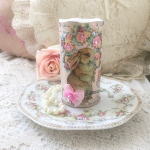 以下が含まれることがあります： ピンクの花とラインストーンで囲まれたウサギが描かれたヴィンテージデザインの白いキャンドル。キャンドルは、花柄の白いプレートと真珠のネックレスの上に置かれています。
