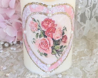 May include: A white pillar candle with a pink and gold heart design featuring a bouquet of pink roses. The candle is decorated with white pearl accents.