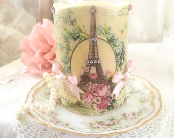 May include: A white candle with a floral design and a picture of the Eiffel Tower. The candle is decorated with pink bows and rhinestones. The candle is sitting on a white saucer with a floral design.
