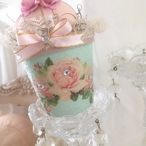 May include: A light blue pincushion with a pink bow and lace trim. The pincushion is decorated with a floral pattern and rhinestones. It is sitting on a clear glass stand with crystal accents.