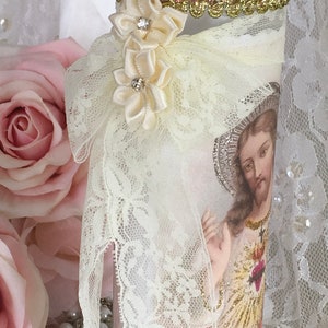 May include: A decorative glass candle holder featuring an image of Jesus. The candle holder is adorned with cream-colored lace, satin ribbon flowers, and a gold and pink jeweled trim around the rim. The background includes pink roses.