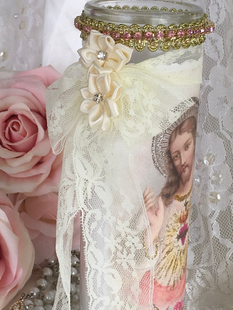 May include: A decorative glass candle holder featuring an image of Jesus. The candle holder is adorned with cream-colored lace, satin ribbon flowers, and a gold and pink jeweled trim around the rim. The background includes pink roses.