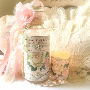 May include: A clear glass bottle with a pink flower and a label that reads "Eau de Cologne &agrave; la Fileuse". The bottle is decorated with rhinestones and pearls. The bottle is sitting on a white lace tablecloth with a candle next to it.