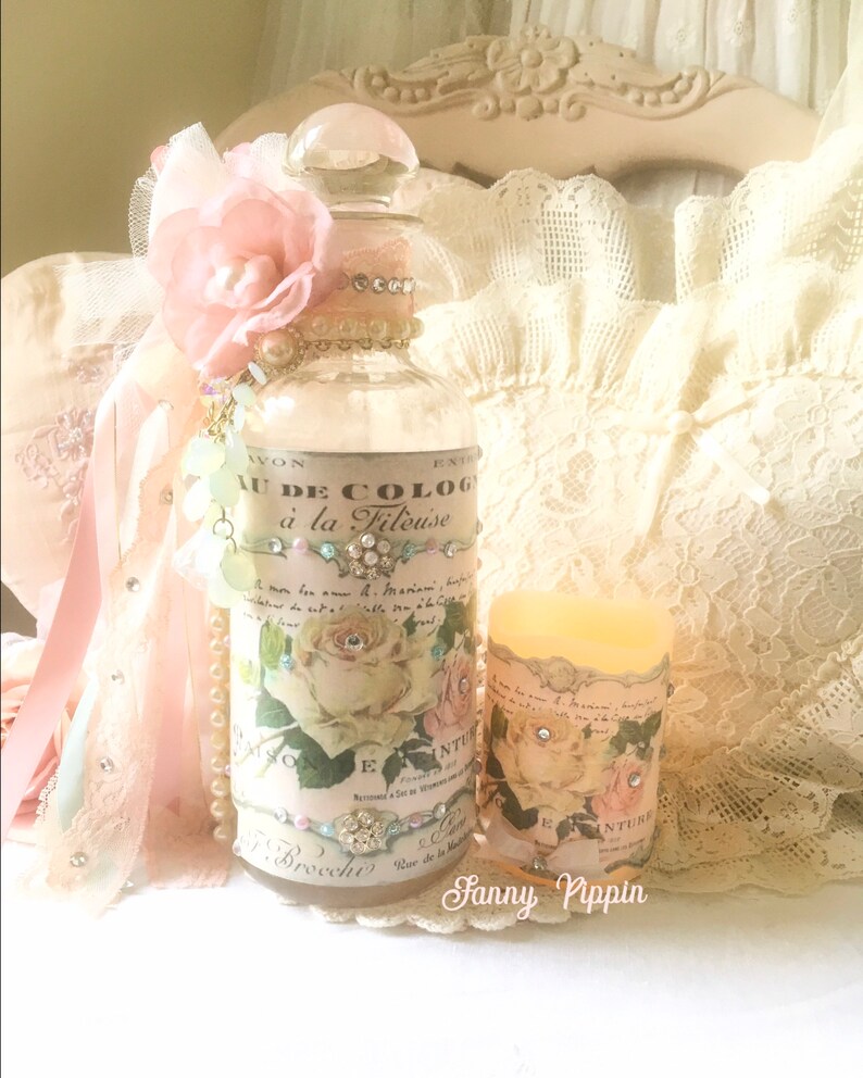 May include: A clear glass bottle with a pink flower and a label that reads "Eau de Cologne &agrave; la Fileuse". The bottle is decorated with rhinestones and pearls. The bottle is sitting on a white lace tablecloth with a candle next to it.