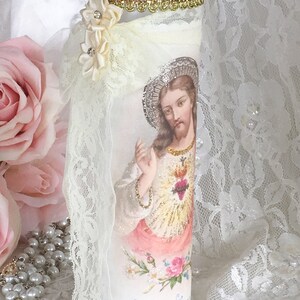 May include: A decorative glass candle holder featuring an image of Jesus Christ. The candle holder is adorned with lace, ribbon, and a pink jeweled trim. A cluster of cream-colored flowers with rhinestones is attached to the side.