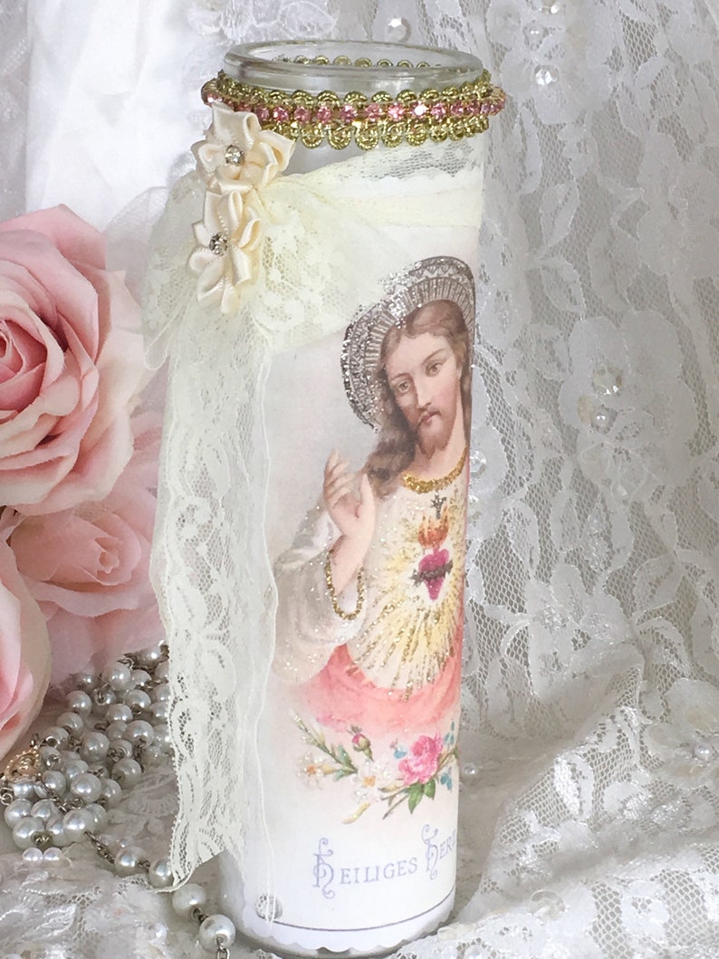 May include: A decorative glass candle holder featuring an image of Jesus Christ. The candle holder is adorned with lace, ribbon, and a pink jeweled trim. A cluster of cream-colored flowers with rhinestones is attached to the side.