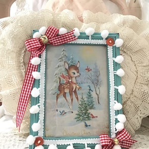 Puede incluir: Un marco de fotos turquesa con una escena navide&ntilde;a vintage con un ciervo, p&aacute;jaros y un paisaje nevado. El marco est&aacute; decorado con pompones blancos, cinta de cuadros rojos y blancos y una peque&ntilde;a campana. Las palabras "Christmas Cheer" son visibles.