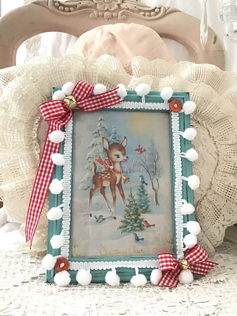 Puede incluir: Un marco de fotos turquesa con una escena navide&ntilde;a vintage con un ciervo, p&aacute;jaros y un paisaje nevado. El marco est&aacute; decorado con pompones blancos, cinta de cuadros rojos y blancos y una peque&ntilde;a campana. Las palabras "Christmas Cheer" son visibles.