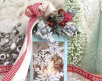 May include: A teal blue lantern with white snowflake cutouts and a white candle inside. The lantern is decorated with a red and white gingham ribbon, a white satin ribbon, and a white lace ribbon. The lantern is surrounded by a white flocked Christmas tree and a white doily.