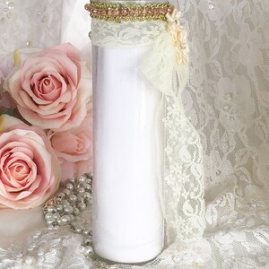 May include: A white glass candle with a decorative gold and pink trim at the top. The candle is adorned with cream-colored lace and a cream-colored bow. Pink roses and a pearl rosary are in the background.