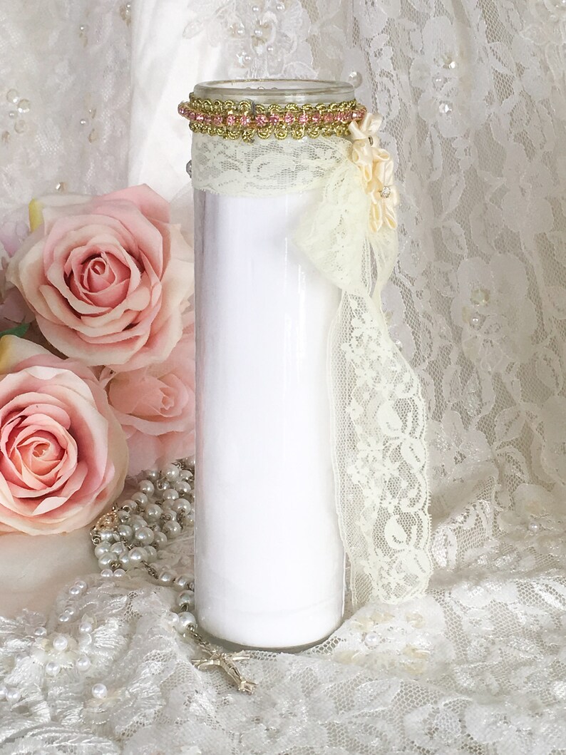 May include: A white glass candle with a decorative gold and pink trim at the top. The candle is adorned with cream-colored lace and a cream-colored bow. Pink roses and a pearl rosary are in the background.