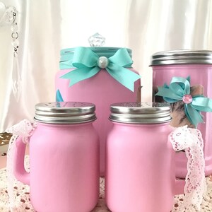 Puede incluir: Conjunto de cuatro tarros de cristal rosa con tapas plateadas. Dos tarros tienen asas y detalles de encaje blanco. Un tarro tiene un lazo turquesa y un pomo de cristal transparente.