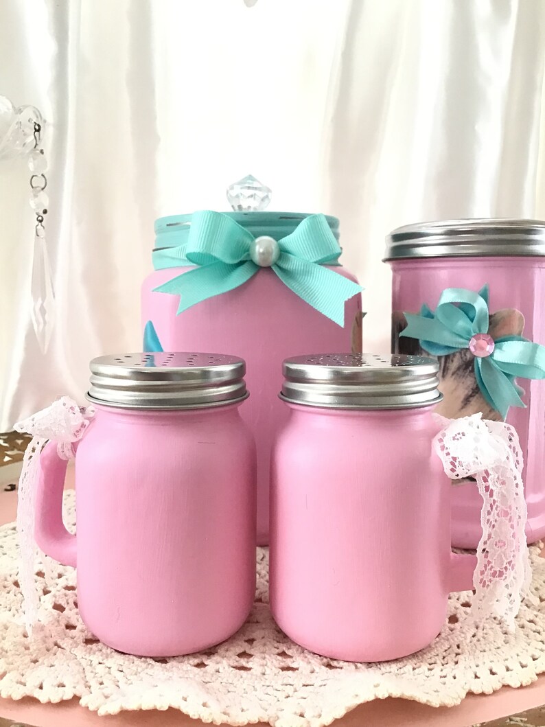 Puede incluir: Conjunto de cuatro tarros de cristal rosa con tapas plateadas. Dos tarros tienen asas y detalles de encaje blanco. Un tarro tiene un lazo turquesa y un pomo de cristal transparente.