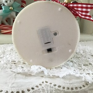 May include: White ceramic Christmas tree ornament with a red and white checkered ribbon and a gold bell. The ornament has a battery compartment on the bottom.
