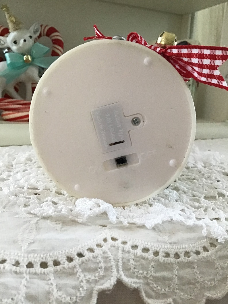 May include: White ceramic Christmas tree ornament with a red and white checkered ribbon and a gold bell. The ornament has a battery compartment on the bottom.