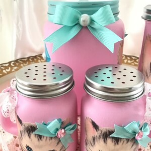 Puede incluir: Un juego de tres tarros de alba&ntilde;il pintados de rosa con un dise&ntilde;o de cara de gatito. Los tarros tienen tapas plateadas y est&aacute;n decorados con lazos turquesa y diamantes de imitaci&oacute;n. El tarro m&aacute;s grande tiene una tapa turquesa con un pomo de cristal transparente.