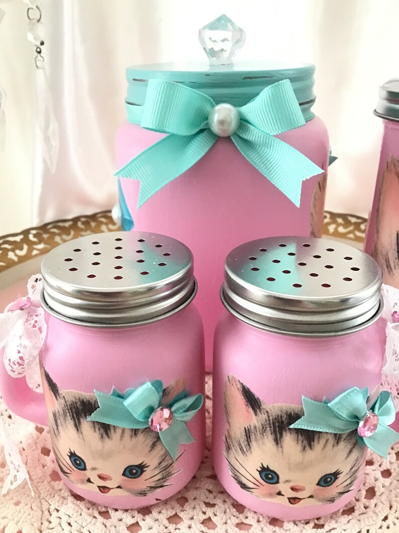 Puede incluir: Un juego de tres tarros de alba&ntilde;il pintados de rosa con un dise&ntilde;o de cara de gatito. Los tarros tienen tapas plateadas y est&aacute;n decorados con lazos turquesa y diamantes de imitaci&oacute;n. El tarro m&aacute;s grande tiene una tapa turquesa con un pomo de cristal transparente.