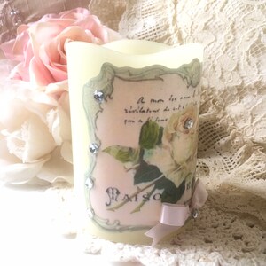 May include: A white pillar candle with a decorative label featuring a floral design and the text "Maison de Fleurs". The label has a green border and is adorned with rhinestones. A pink ribbon is tied around the bottom of the candle.