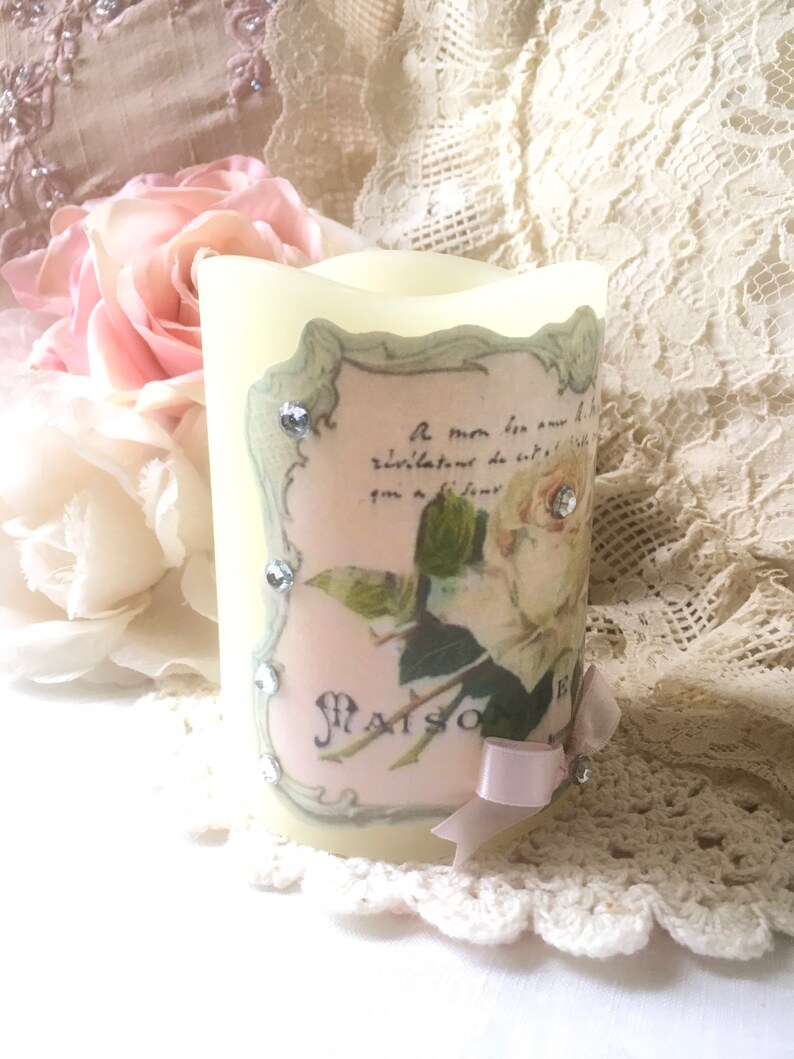May include: A white pillar candle with a decorative label featuring a floral design and the text "Maison de Fleurs". The label has a green border and is adorned with rhinestones. A pink ribbon is tied around the bottom of the candle.