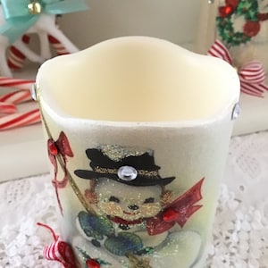 May include: White candle with a vintage Christmas image of a snowman holding a broom. The snowman is wearing a black hat with a gold band and a red scarf. The candle has a red and white ribbon tied around the bottom.