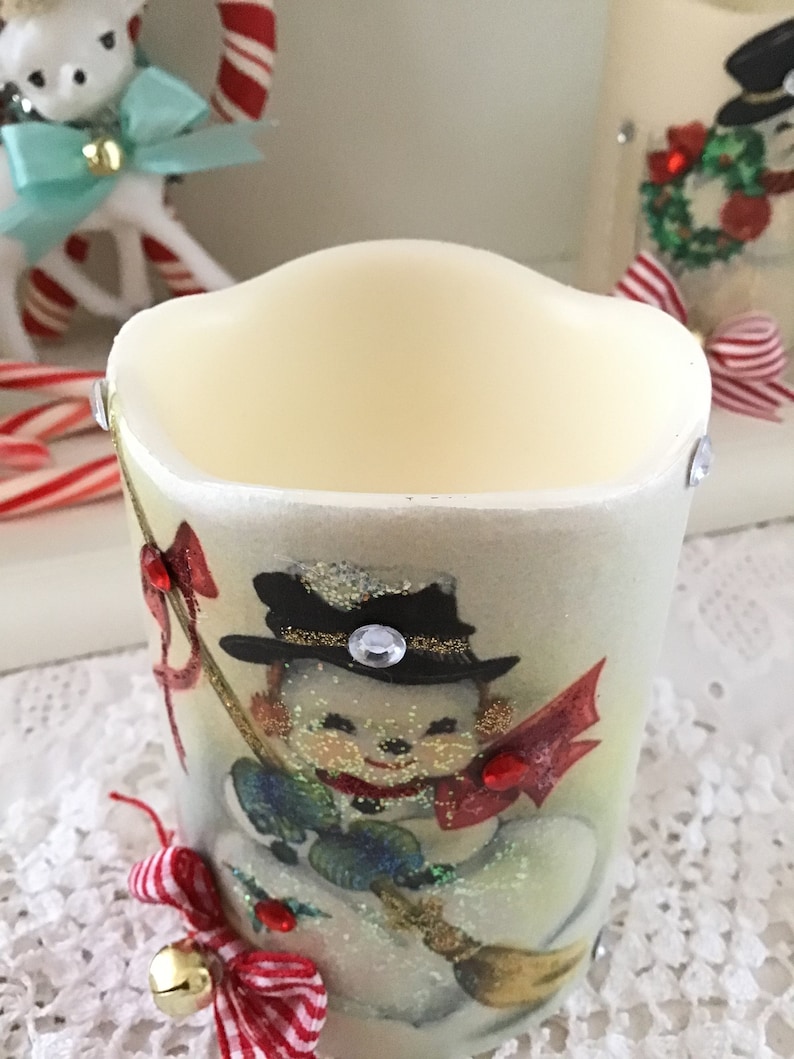 May include: White candle with a vintage Christmas image of a snowman holding a broom. The snowman is wearing a black hat with a gold band and a red scarf. The candle has a red and white ribbon tied around the bottom.