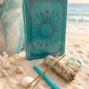 May include: A teal journal featuring a sun and moon phase design on the cover, accompanied by a teal pen, a bundle of dried sage, two incense sticks, a small round candle with embedded seashells, and three loose seashells arranged on white sand.